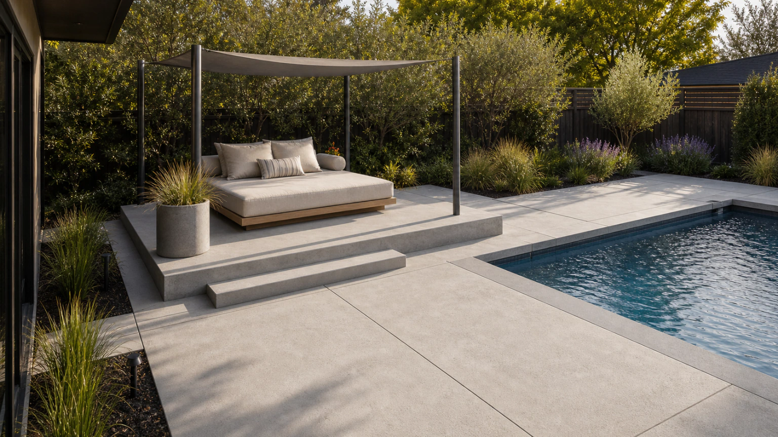 Concrete pool deck with a raised daybed platform, shade canopy, and low privacy planting.